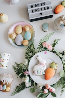 An arrangement of Easter-themed items on a white surface, including decorated cookies shaped like a bunny and a carrot placed on a white plate surrounded by green leaves and pink flowers. There are pastel-colored eggs on a dish, a cup with colorful polka dots, a mini carrot decoration, and a lit-up sign with the text 'HAPPY EASTER' and a heart symbol.