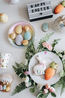 An arrangement of Easter-themed items on a white surface, including decorated cookies shaped like a bunny and a carrot placed on a white plate surrounded by green leaves and pink flowers. There are pastel-colored eggs on a dish, a cup with colorful polka dots, a mini carrot decoration, and a lit-up sign with the text 'HAPPY EASTER' and a heart symbol.