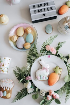 An arrangement of Easter-themed items on a white surface, including decorated cookies shaped like a bunny and a carrot placed on a white plate surrounded by green leaves and pink flowers. There are pastel-colored eggs on a dish, a cup with colorful polka dots, a mini carrot decoration, and a lit-up sign with the text 'HAPPY EASTER' and a heart symbol.