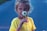 A young girl is holding a white daisy up to her face. She wears a bright yellow shirt, and her hair is loosely tied back. The background is muted and slightly out of focus, emphasizing her and the flower.
