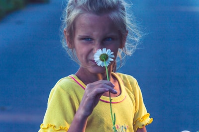 A young girl is holding a white daisy up to her face. She wears a bright yellow shirt, and her hair is loosely tied back. The background is muted and slightly out of focus, emphasizing her and the flower.