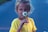 A young girl is holding a white daisy up to her face. She wears a bright yellow shirt, and her hair is loosely tied back. The background is muted and slightly out of focus, emphasizing her and the flower.
