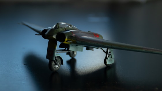 A small, detailed model of a vintage military aircraft is positioned on a dark surface. The aircraft features a distinctive design with straight wings and a sleek fuselage. The lighting creates a dramatic shadow underneath, highlighting the model’s intricate details.
