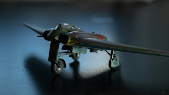 A small, detailed model of a vintage military aircraft is positioned on a dark surface. The aircraft features a distinctive design with straight wings and a sleek fuselage. The lighting creates a dramatic shadow underneath, highlighting the model’s intricate details.