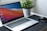 A laptop with a colorful abstract wallpaper on the screen is placed on a dark mat. To the right of the laptop, a wireless mouse is positioned. In the background, a green potted plant in a textured white pot adds a touch of nature to the desk setup.