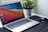 A laptop with a colorful abstract wallpaper on the screen is placed on a dark mat. To the right of the laptop, a wireless mouse is positioned. In the background, a green potted plant in a textured white pot adds a touch of nature to the desk setup.
