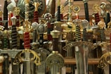 A collection of ornate swords with elaborately designed hilts and scabbards. The swords showcase various intricate details with inlays, engravings, and symbols, many adorned with gold, silver, and colored handles. They are positioned vertically, densely packed together, and create a visually striking display.