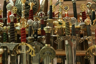 A collection of ornate swords with elaborately designed hilts and scabbards. The swords showcase various intricate details with inlays, engravings, and symbols, many adorned with gold, silver, and colored handles. They are positioned vertically, densely packed together, and create a visually striking display.