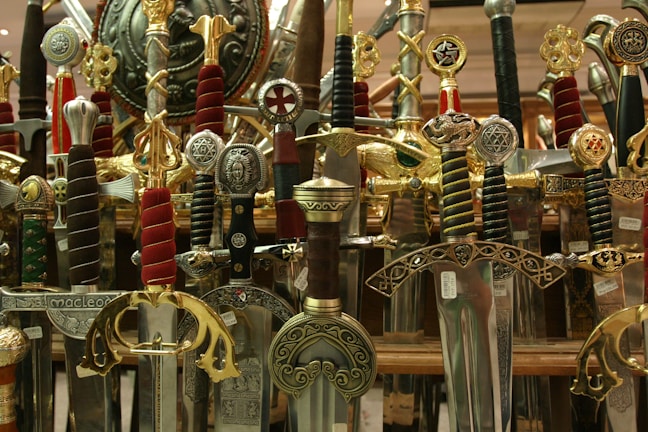 A collection of ornate swords with elaborately designed hilts and scabbards. The swords showcase various intricate details with inlays, engravings, and symbols, many adorned with gold, silver, and colored handles. They are positioned vertically, densely packed together, and create a visually striking display.