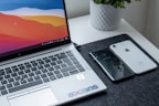 A laptop with a colorful screen is placed on a table. To the right of the laptop, two smartphones, one black and one white, are lying next to each other. A potted plant can be seen in the background. The table has a dark mat beneath the laptop and phones, and the keyboard of the laptop includes a sticker with a logo on it.