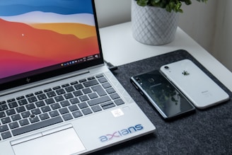 A laptop with a colorful screen is placed on a table. To the right of the laptop, two smartphones, one black and one white, are lying next to each other. A potted plant can be seen in the background. The table has a dark mat beneath the laptop and phones, and the keyboard of the laptop includes a sticker with a logo on it.