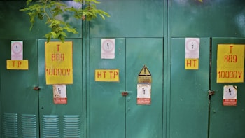 A row of green industrial electrical cabinets with various warning labels attached, including yellow signs with red lettering indicating high voltage. A small tree branch with green leaves is visible along the top edge.