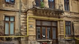 An old, weathered building with deteriorating plaster and exposed brick. Decorative elements like iron railings and detailed moldings are present near the windows and balcony. A yellow gas pipe runs along the facade, adding a modern element to the otherwise historic architecture. Some windows display peeling paint, contributing to the overall worn look.