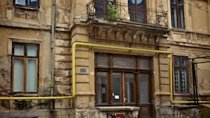 An old, weathered building with deteriorating plaster and exposed brick. Decorative elements like iron railings and detailed moldings are present near the windows and balcony. A yellow gas pipe runs along the facade, adding a modern element to the otherwise historic architecture. Some windows display peeling paint, contributing to the overall worn look.