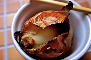 A roasted onion with slightly charred, crispy outer layers sits in a small white bowl. Chopsticks rest on the edge of the bowl. The background consists of a tiled surface.