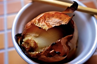 A roasted onion with slightly charred, crispy outer layers sits in a small white bowl. Chopsticks rest on the edge of the bowl. The background consists of a tiled surface.
