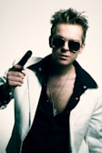 A person with spiky hair wearing dark sunglasses and a black shirt under a white suit jacket holds a gun. A necklace with a dog tag hangs around their neck.