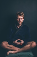 A person is sitting cross-legged with closed eyes, exhibiting a meditative posture. The setting is dimly lit, highlighting the contemplative mood. The individual is wearing a dark robe and is positioned against a plain, dark background.