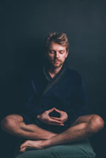 A person is sitting cross-legged with closed eyes, exhibiting a meditative posture. The setting is dimly lit, highlighting the contemplative mood. The individual is wearing a dark robe and is positioned against a plain, dark background.