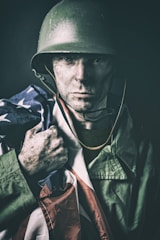 A soldier with dirt-smeared face and intense expression holds an American flag draped over his shoulder. He wears a military helmet and rugged uniform, embodying a sense of determination and resolve.