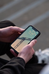 A person holds a smartphone displaying a lock screen with an image of a construction crane against a pale sky. The phone shows the time and date, indicating it is the evening. The individual is sitting down, wearing dark clothing, and their hands are visible.