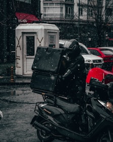A motorcycle delivery person is riding through a snowy urban street, dressed in dark, weather-proof clothing and carrying a large delivery box. Snowflakes are gently falling, and the ground is wet and reflective. In the background, there are parked cars, a white booth with graffiti, and buildings with shop signs.