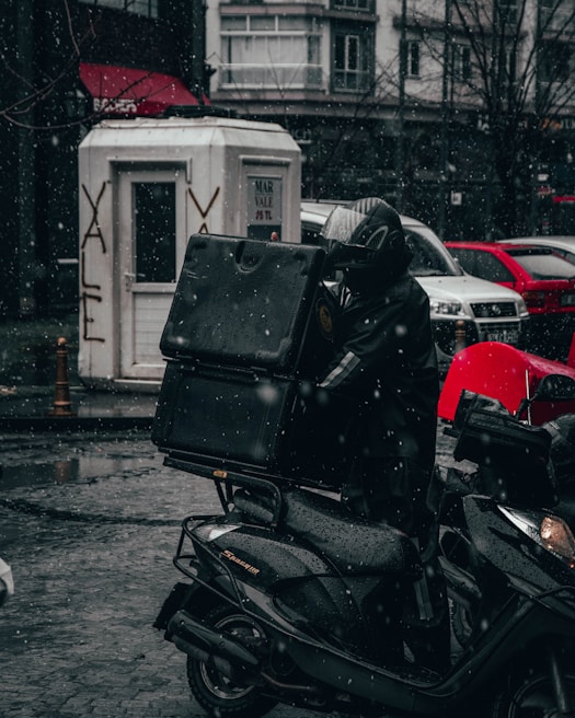 A motorcycle delivery person is riding through a snowy urban street, dressed in dark, weather-proof clothing and carrying a large delivery box. Snowflakes are gently falling, and the ground is wet and reflective. In the background, there are parked cars, a white booth with graffiti, and buildings with shop signs.