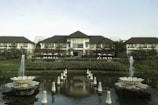 A grand resort-style building is set amidst lush greenery and features a large, symmetrical garden with two decorative fountains in the foreground. The building has a multi-peaked roofline and is flanked by trees and manicured shrubs. A pond with ornate sculptures is reflected in the calm water.