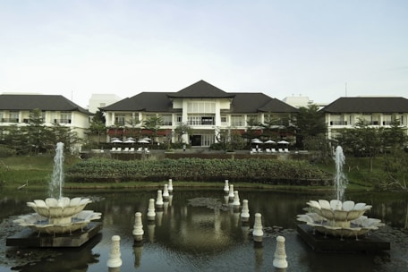 A grand resort-style building is set amidst lush greenery and features a large, symmetrical garden with two decorative fountains in the foreground. The building has a multi-peaked roofline and is flanked by trees and manicured shrubs. A pond with ornate sculptures is reflected in the calm water.