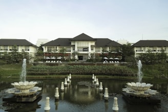 A grand resort-style building is set amidst lush greenery and features a large, symmetrical garden with two decorative fountains in the foreground. The building has a multi-peaked roofline and is flanked by trees and manicured shrubs. A pond with ornate sculptures is reflected in the calm water.