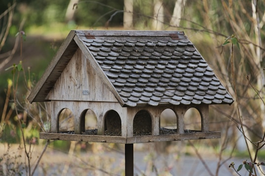 A wooden birdhouse with a shingled roof is mounted on a post. It features several arched openings and is positioned in a natural setting with blurred background foliage. The surrounding branches and leaves add to the rustic and serene atmosphere.