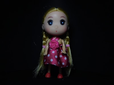 A doll with large, wide black eyes and long blonde hair styled in braids. It is wearing a pink polka dot dress with a large flower on the front, and has matching pink shoes. The background is black, creating a dark contrast with the brightly colored doll.
