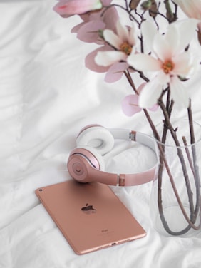 A pair of rose gold headphones is placed next to a matching iPad on a white fabric surface. Beside them, a vase holds several branches with large, delicate white and pink flowers, adding a touch of elegance to the scene.