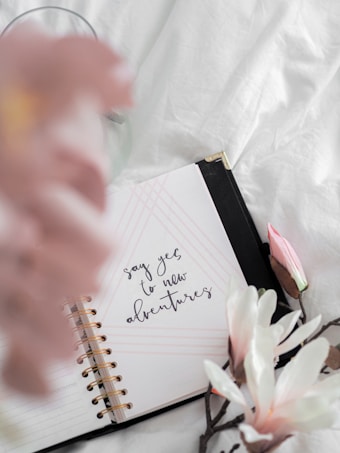 An open notebook with a spiral binding rests on a soft white surface. The notebook displays the phrase 'say yes to new adventures' written in a stylish, cursive font. Surrounding the notebook are soft pink and white flowers, which add a touch of elegance and calmness to the composition.