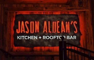 A glowing neon sign reads 'Jason Aldean's Kitchen + Rooftop Bar' set against a rustic wood background. The words 'Jason Aldean's' are in bold red neon, while 'Kitchen + Rooftop Bar' is in bright white. A metal railing is visible in the lower right corner.