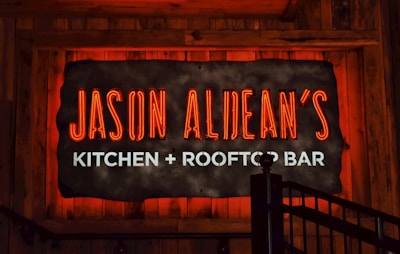 A glowing neon sign reads 'Jason Aldean's Kitchen + Rooftop Bar' set against a rustic wood background. The words 'Jason Aldean's' are in bold red neon, while 'Kitchen + Rooftop Bar' is in bright white. A metal railing is visible in the lower right corner.