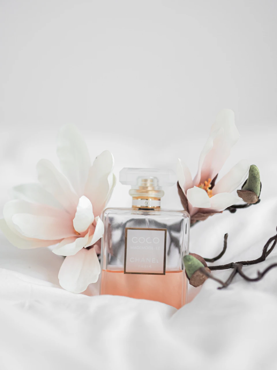 The image presents a bottle of perfume, specifically Chanel No. 5, resting on a white bedspread with two large white flowers positioned to its left and right sides. The perfume bottle is placed centrally within the frame, drawing attention to it as the main subject of the composition.