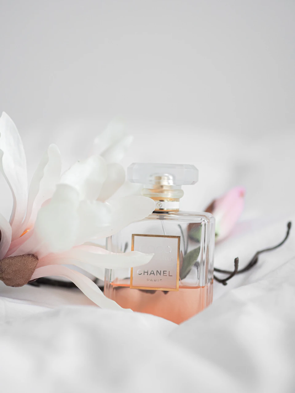 The image presents a scene featuring a bottle of Chanel perfume, a white flower with pink petals, and some black and brown sticks arranged on a bedspread or white sheet. The perfume bottle is positioned centrally within the frame, while the flower and sticks are located to its left and right sides respectively.

The background appears to be a plain white surface, which contrasts with the vibrant colors of the perfume bottle and the delicate petals of the flower.