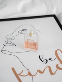 A perfume bottle is placed on a sheet of paper with line art of a face and the words 'be kind' printed in a cursive font. The paper is framed, giving it an artistic and elegant presentation.