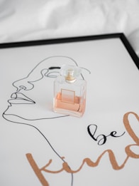 A perfume bottle is placed on a sheet of paper with line art of a face and the words 'be kind' printed in a cursive font. The paper is framed, giving it an artistic and elegant presentation.