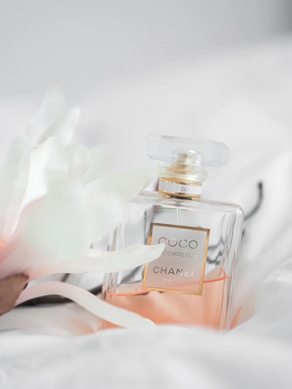The image features a bottle of Chanel perfume resting on a white bedspread, with its label facing upwards and the perfume bottle positioned centrally within the frame. The background is blurred, drawing attention to the perfume bottle. A single flower can be seen in the top left corner of the image, adding a touch of nature's beauty to the scene.

The perfume bottle itself has a white body adorned with gold accents, giving it an elegant and sophisticated appearance. The label on the bottle displays the Chanel logo, indicating that this is a Chanel perfume.