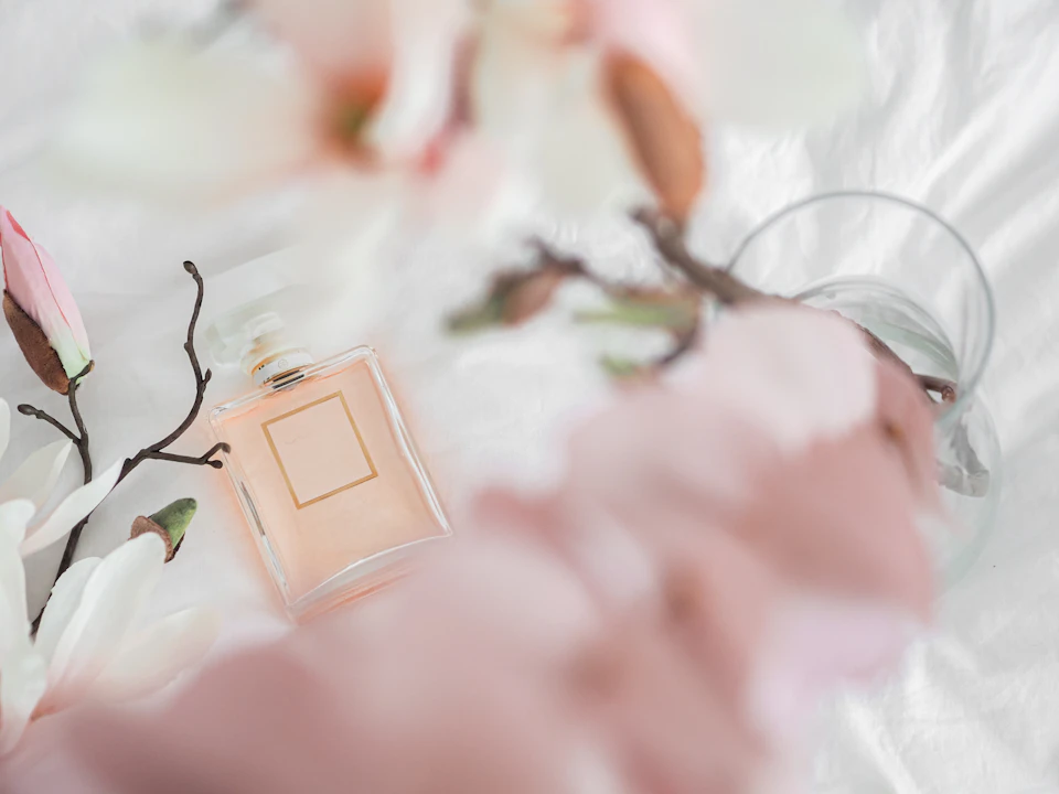 The image presents a scene set on a bed with white sheets and a pink floral comforter, creating an elegant and romantic atmosphere. In the center of the image is a bottle of perfume, its label clearly visible against the soft hues of the background. The perfume bottle has a gold-colored cap that gleams subtly under the light. Surrounding the perfume bottle are various flowers in different stages of bloom, adding a touch of nature's beauty to the scene.

In addition to the perfume and flowers, there is also a glass vase filled with water placed on the bed, contributing to the overall ambiance of relaxation and tranquility.
