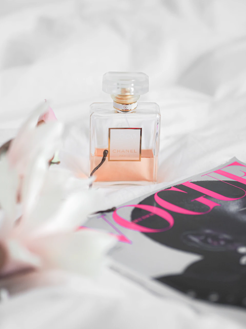 The image presents a scene set on a bed with a white comforter and a pink flower resting on it. In the center of the bed, there is an open perfume bottle with a clear glass body and a gold cap, which appears to be a Chanel perfume. The perfume bottle is positioned in front of a magazine cover featuring a black cat.