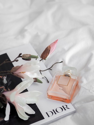 A perfume bottle lies atop a magazine, surrounded by delicate white flowers on a soft white fabric background. The bottle is rectangular with a peach-colored liquid and a golden label. The magazine is predominantly white, featuring the word 'Dior' prominently in bold letters.