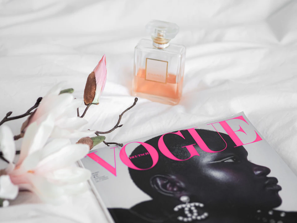 The image presents a scene on a bed with a white comforter, featuring an open magazine titled "Vogue" lying flat on its side. The magazine is adorned with a pink cover and displays an image of a woman's face wearing a necklace. To the left of the magazine, there are two flowers - one white and one pink in color. A bottle of perfume, specifically Chanel No. 5, is also present on the bed.