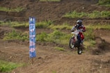 A person riding a dirt bike performs a small jump on a rugged outdoor track. The rider is wearing protective gear, including a helmet and colorful clothing. To the left, there is a vertical banner that displays event information. The background is a mix of dirt tracks and patches of green vegetation.