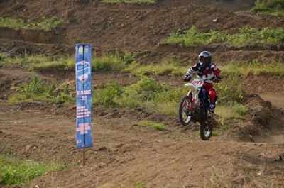 A person riding a dirt bike performs a small jump on a rugged outdoor track. The rider is wearing protective gear, including a helmet and colorful clothing. To the left, there is a vertical banner that displays event information. The background is a mix of dirt tracks and patches of green vegetation.