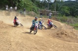 Several motorcyclists race on a dirt track surrounded by lush greenery and trees. Each rider is wearing colorful protective gear and helmets. Dust rises behind the bikes, indicating movement and speed. Spectators watch from the side of the track.