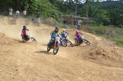 Several motorcyclists race on a dirt track surrounded by lush greenery and trees. Each rider is wearing colorful protective gear and helmets. Dust rises behind the bikes, indicating movement and speed. Spectators watch from the side of the track.