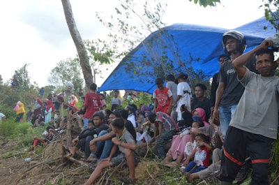 A group of people gather under and around a large blue tarp in an outdoor setting. Some are sitting on the ground or on logs, while others stand. There is a mix of men, women, and children wearing casual clothing, with some individuals sporting hats or helmets. Surrounding the group are trees and natural foliage, suggesting a rural or wilderness location.
